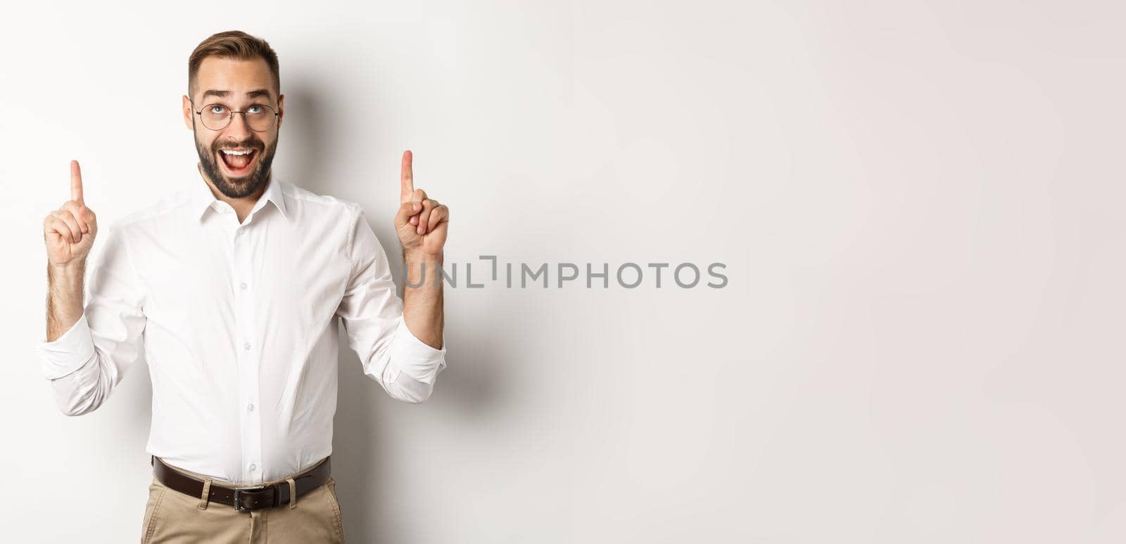Excited businessman checking out advertisement, pointing and looking up with happy face, standing against white background.
