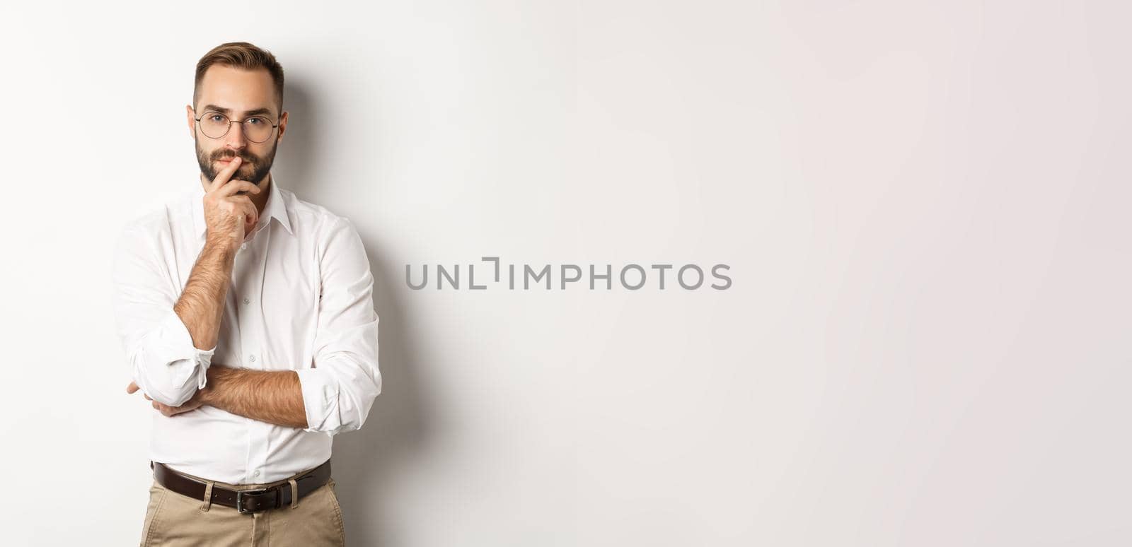 Thoughtful handsome businessman looking at camera, making choice or thinking, standing in glasses and white collar shirt against studio background.