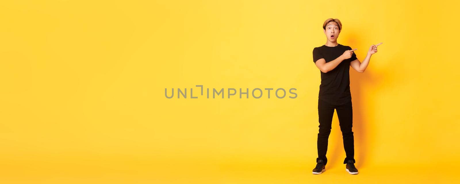 Full-length of amazed and impressed attractive asian guy pointing fingers upper right corner, showing logo, yellow background by Benzoix