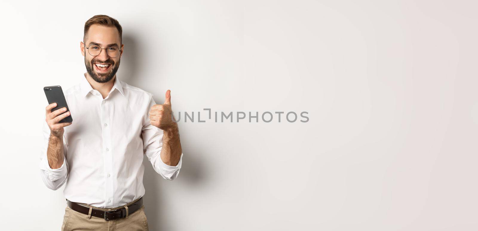 Satisfied business man showing thumbs up after using mobile phone, standing pleased over white background.