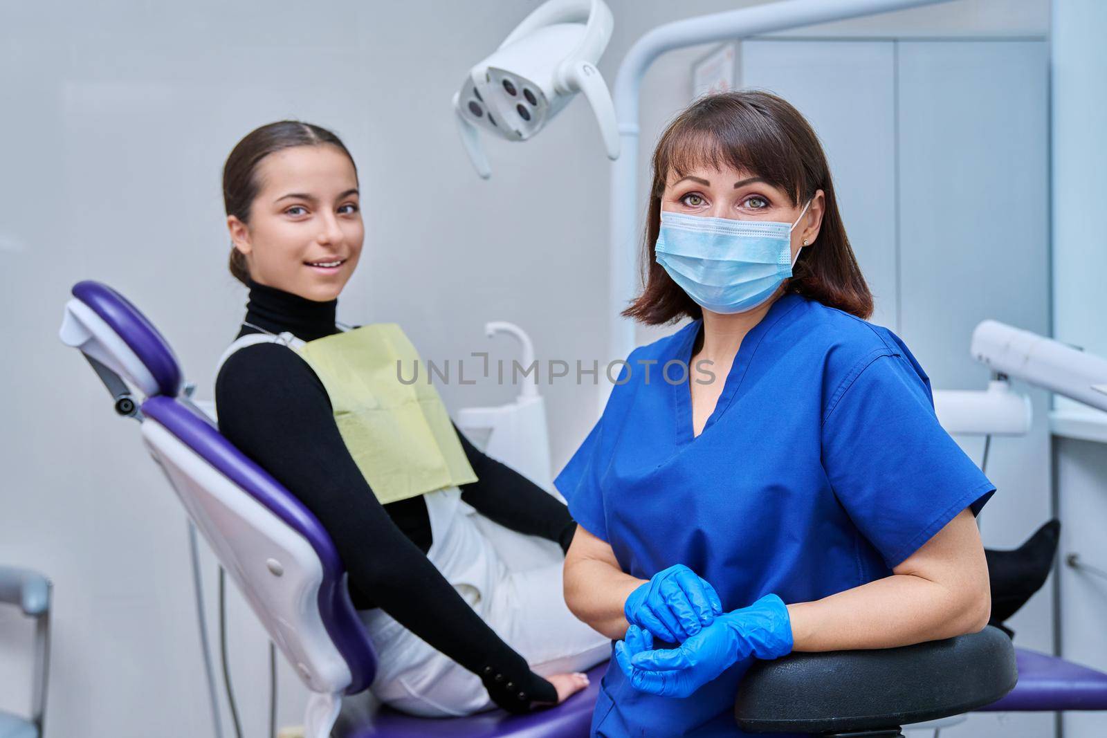 Portrait of female dentist looking at camera with young teenage girl patient sitting in dental chair. Dentistry, hygiene, treatment, dental health care concept