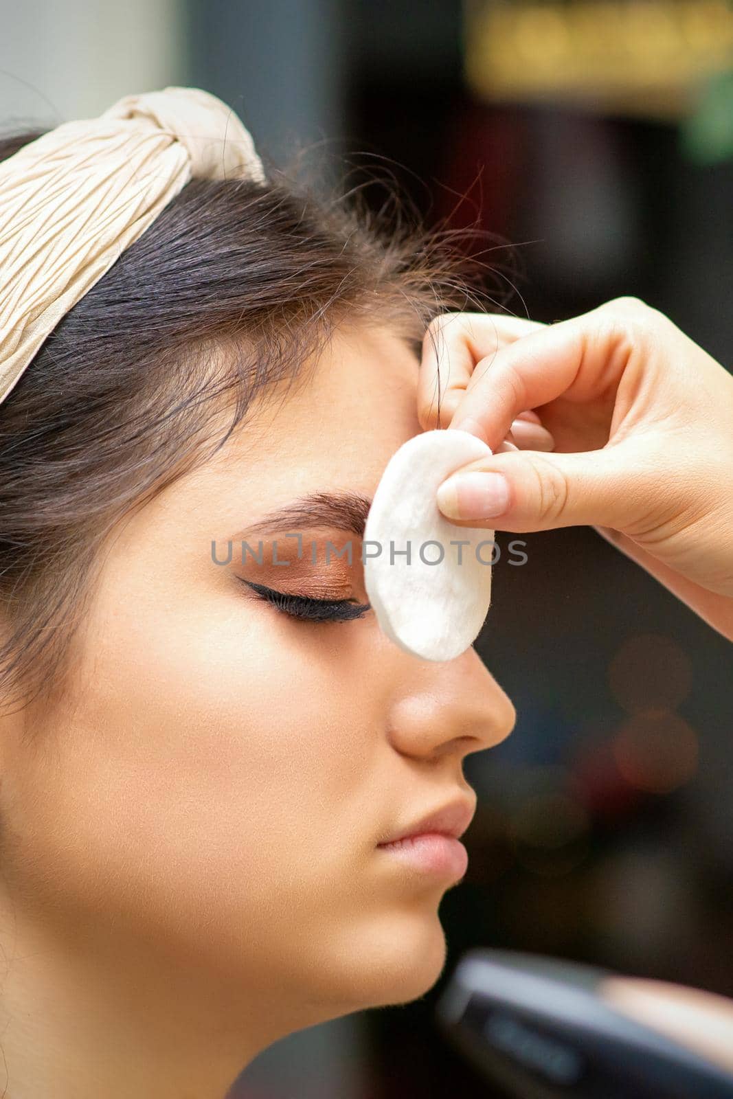 Side view of a makeup artist covers female eye with a cotton pad using airbrush making makeup foundation on her face in a beauty salon