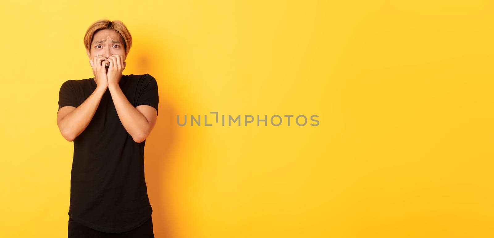 Portrait of scared insecure asian blond guy, holding hands over mouth horrified, looking frightened, yellow background.