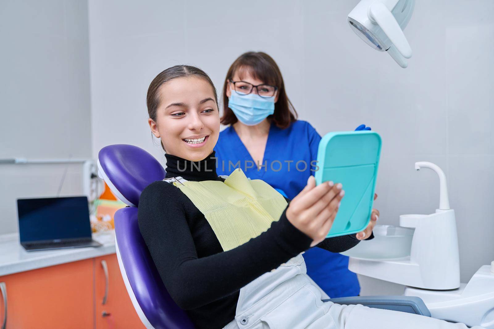 Happy young teenage female with mirror sitting in dentist chair smiling looking at healthy teeth, with dentist doctor in office. Dentistry, hygiene, treatment, dental health care concept