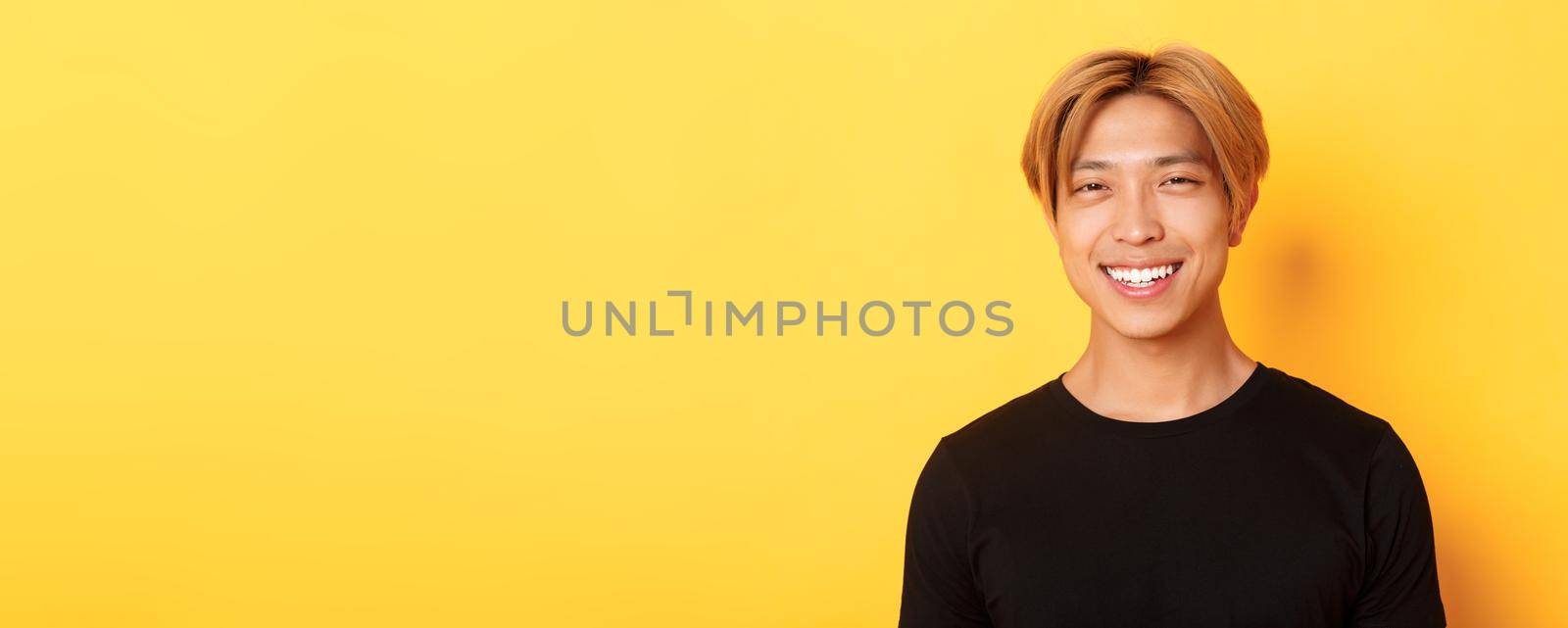 Close-up of handsome stylish asian guy with fair hair, smiling happy over yellow background by Benzoix