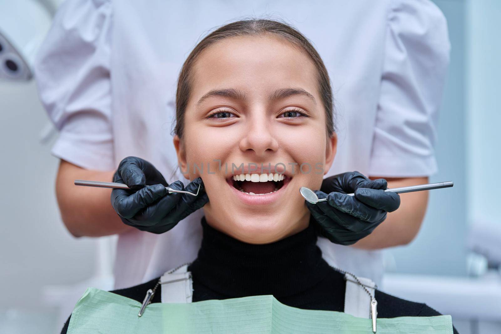 Portrait of young teenage girl in dental chair with hands of doctor with tools. Female teenager smiling with teeth looking at camera in dentist office. Adolescence hygiene treatment dental health care