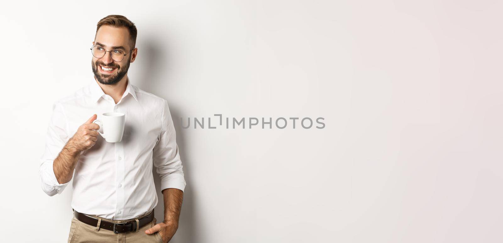 Successful business man drinking coffee, looking sideways with satisfied smile, standing over white background.