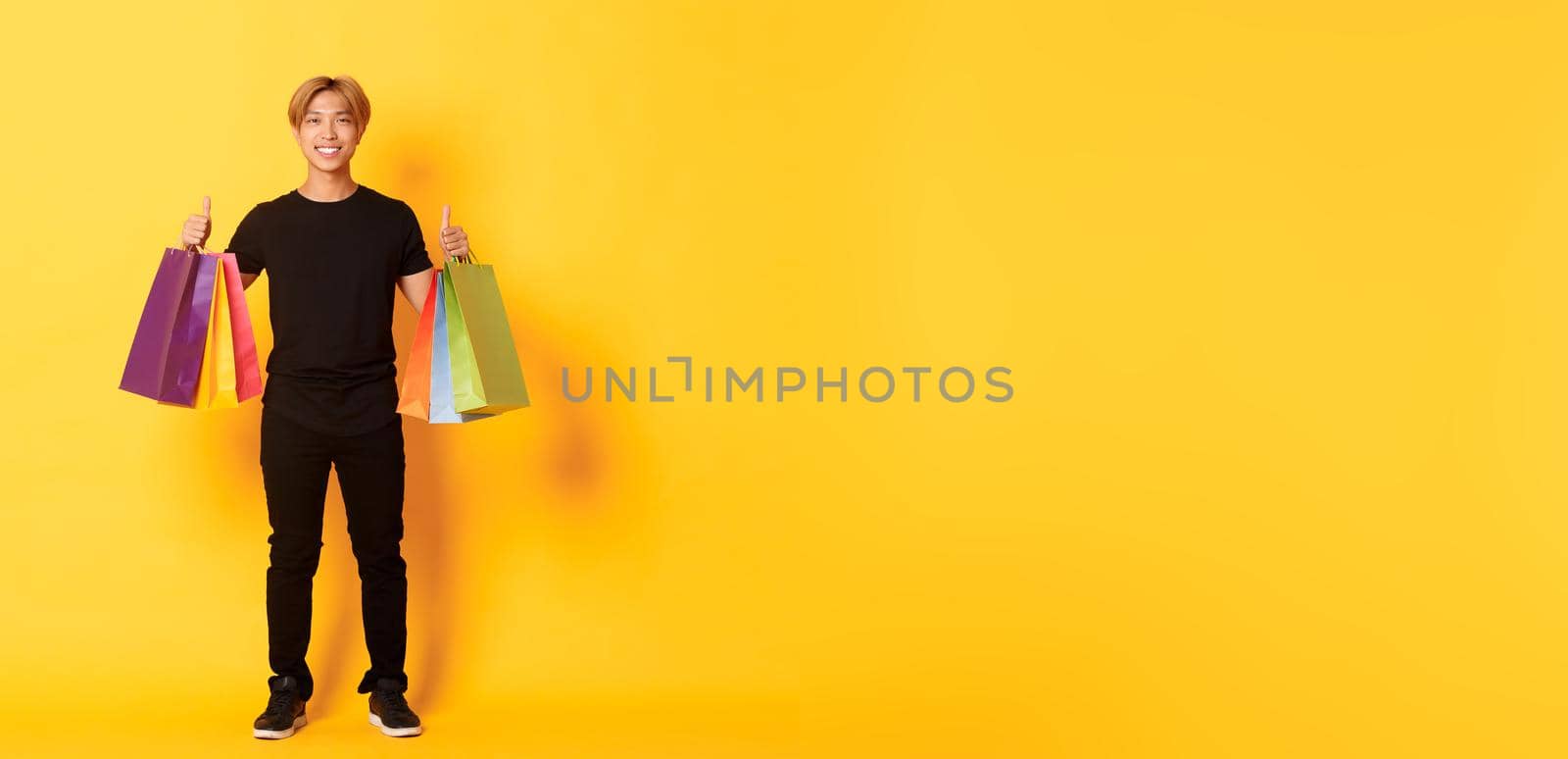 Full-length of attractive korean guy in black clothes, showing thumbs-up, holding shopping bags, yellow background.