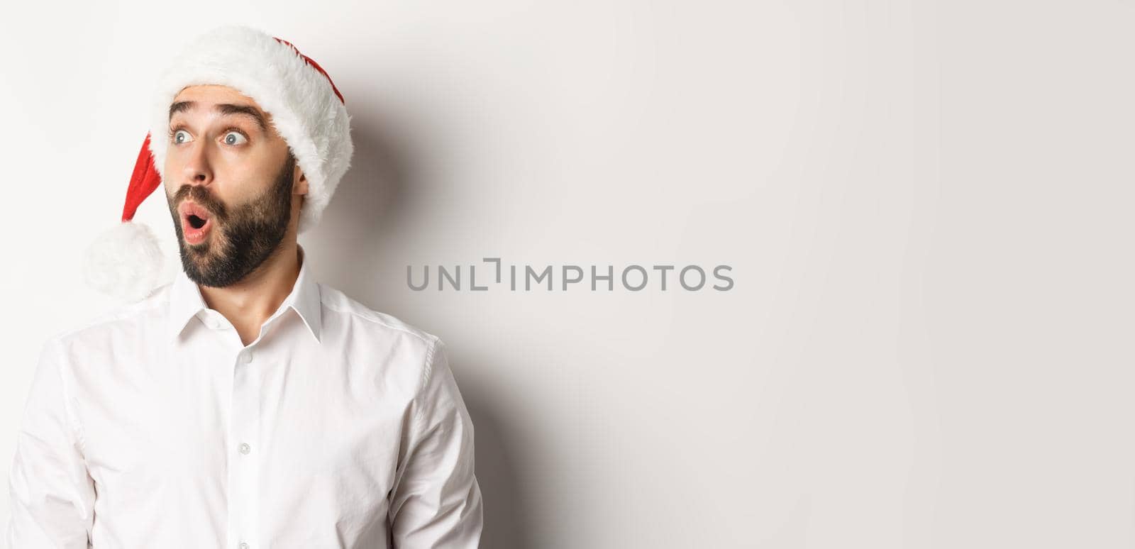 Close-up of amazed bearded man in santa hat, looking left with surprised face, christmas promo offer, standing over white background.