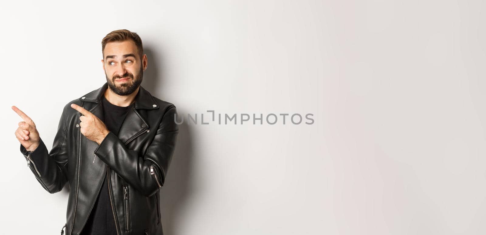 Skeptical and doubtful guy in black leather jacket, shrugging while pointing at upper left corner promo offer, standing over white background.