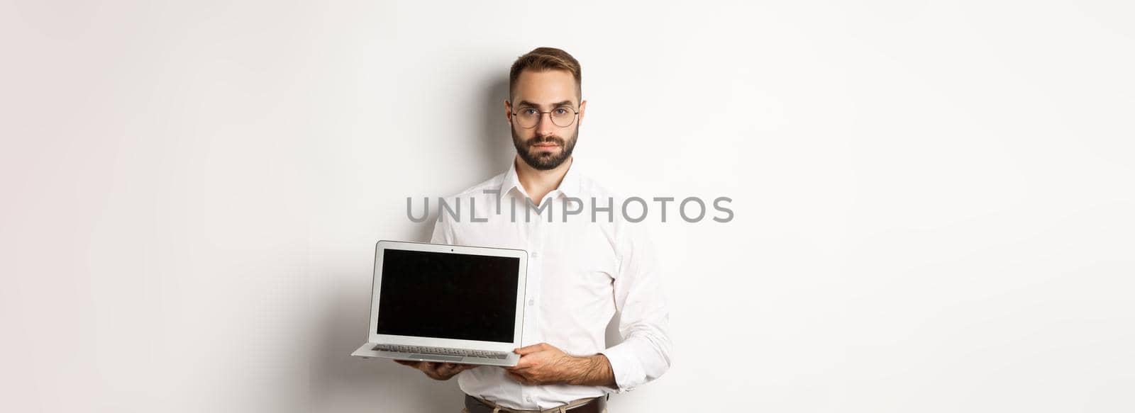 Confident manager demonstrating presentation on screen, showing laptop display and looking serious, standing over white background.
