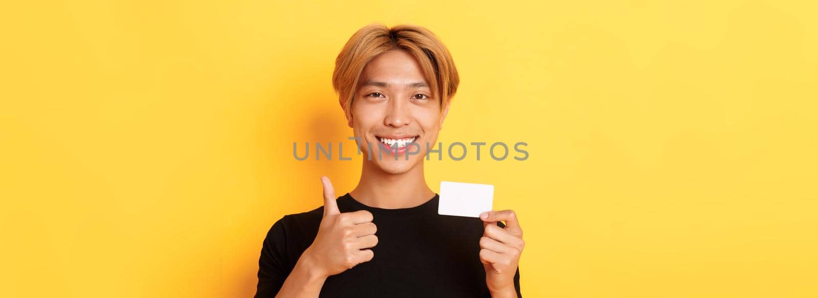 Close-up of satisfied asian guy recommend bank, showing credit card and standing over yellow background.