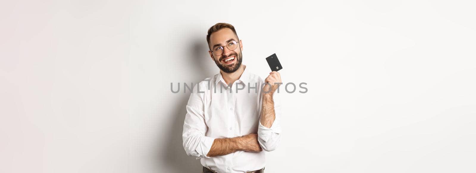 Image of handsome man thinking about shopping and holding credit card, looking at upper left corner thoughtful, white background.