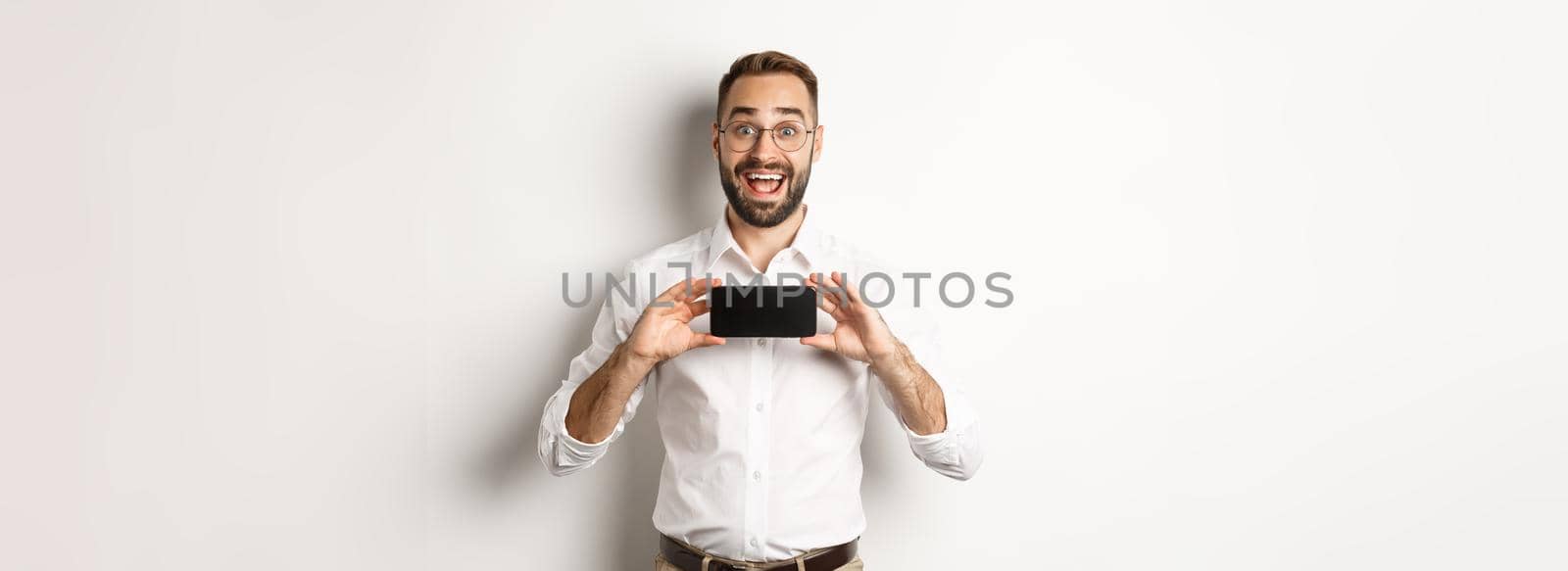 Excited handsome man showing mobile phone screen, looking amazed, standing over white background.