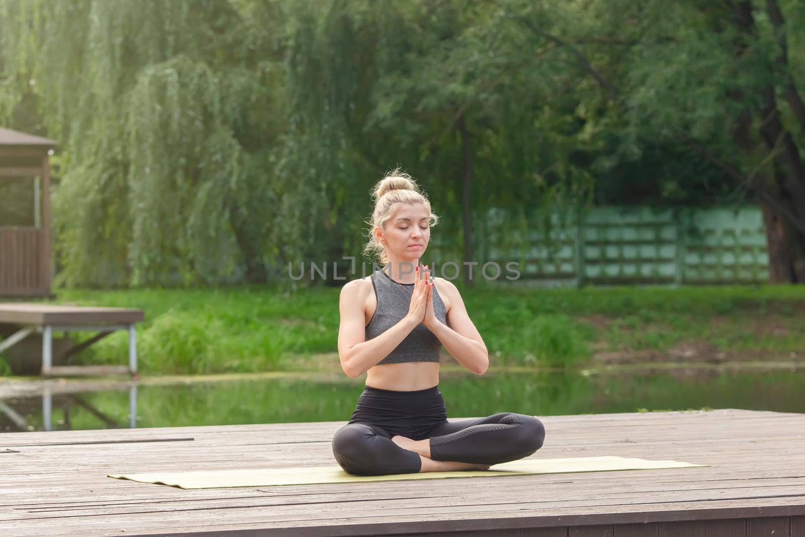 A beautiful woman in a gray top and leggings, sitting on a wooden platform by a pond in the park in summer, does yoga with her palms together, closing her eyes