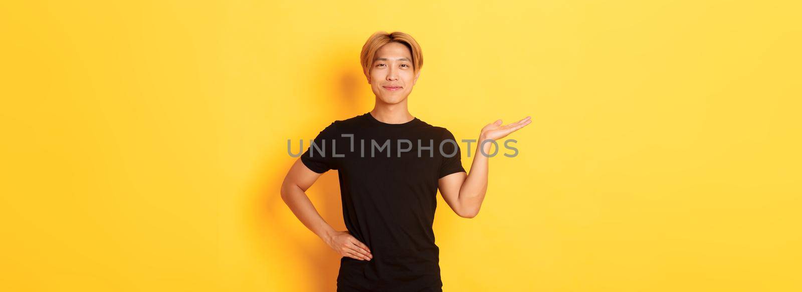 Portrait of happy and proud smiling asian guy holding something on hand over yellow background.
