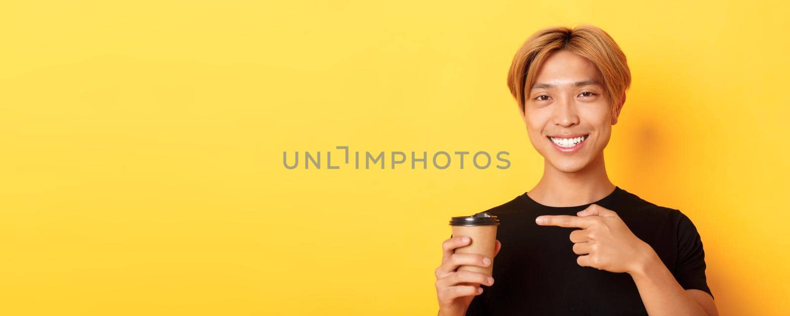 Close-up of satisfied handsome asian blond guy, smiling delighted and pointing finger at delicious cup of coffee, standing over yellow background.