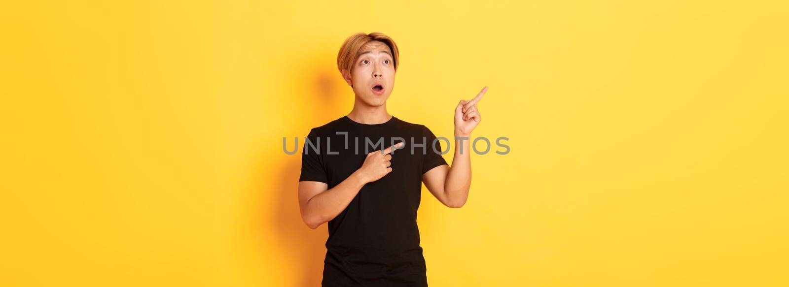 Portrait of impressed asian guy looking and pointing fingers upper left corner, standing yellow background.