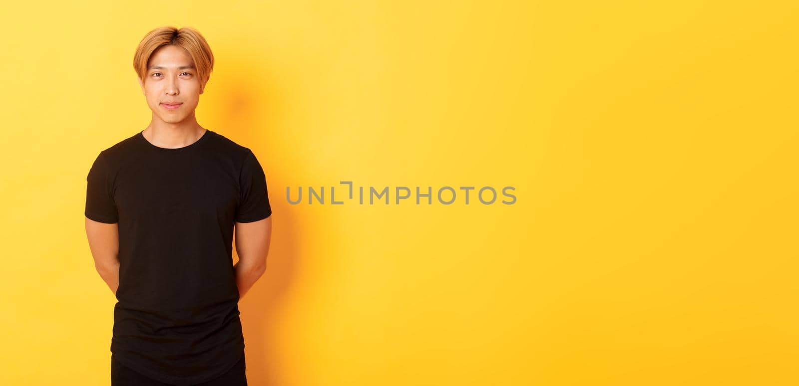 Portrait of handsome smiling asian blond guy looking sassy, holding hands behind back, standing yellow background.