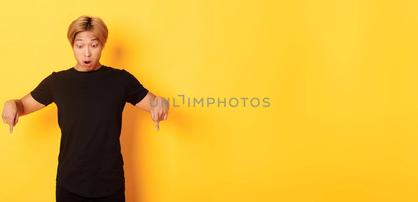 Portrait of surpirsed asian guy gasping amazed, pointing fingers and looking down at banner, standing yellow background by Benzoix