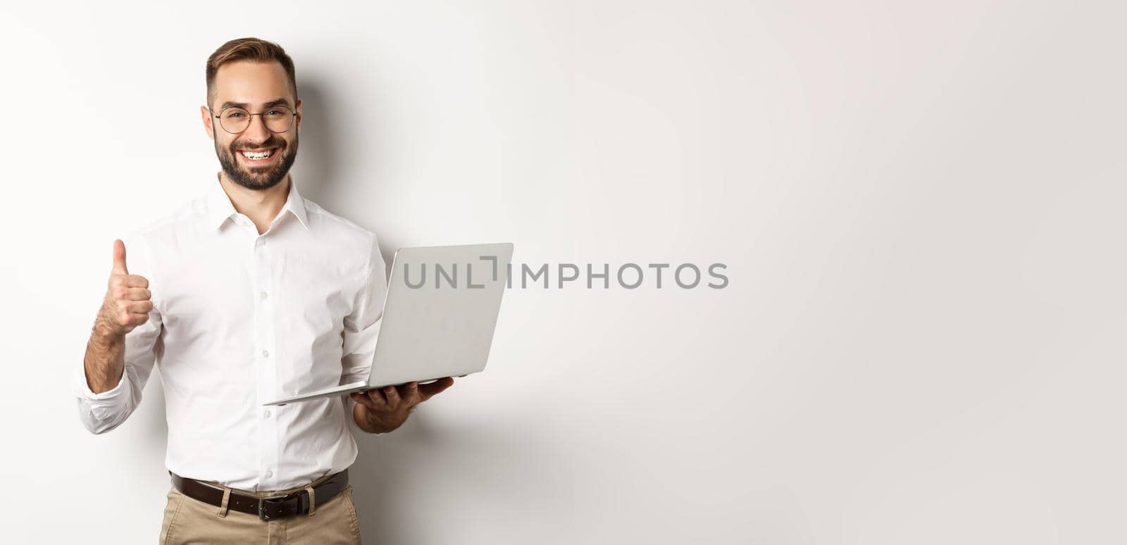 Business. Young successful male entrepreneur showing thumb-up while working on laptop, standing over white background.