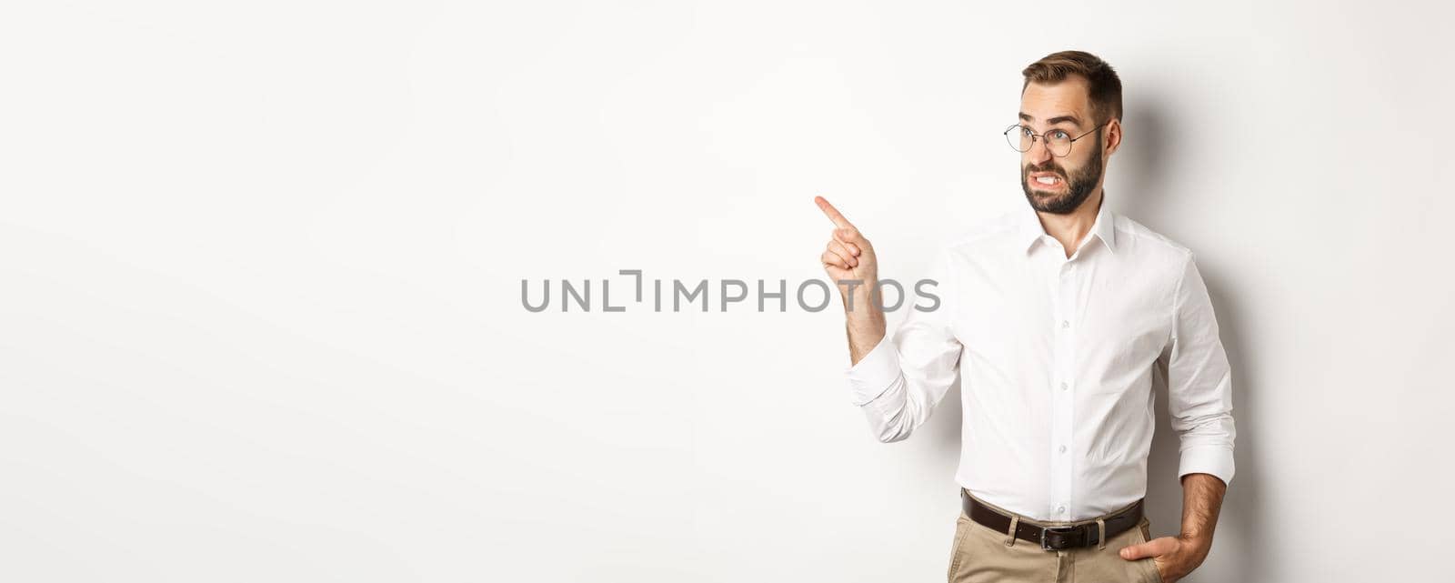 Young bearded guy seeing something disturbing, cringe while pointing finger left at promo offer, standing awkward against white background.