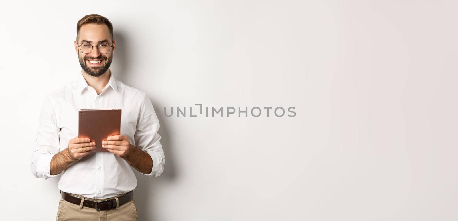 Handsome bearded man in glasses using digital tablet, smiling satisfied, standing against white background by Benzoix