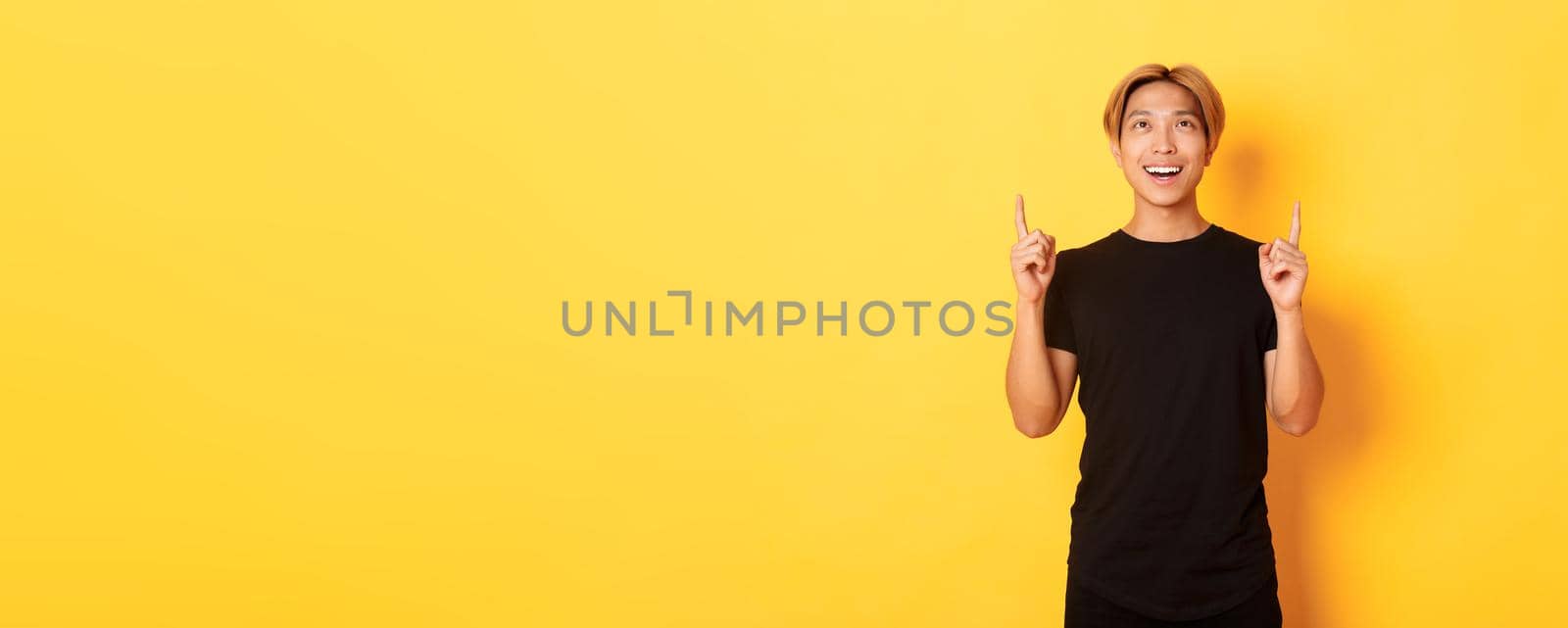 Portrait of satisfied asian blond guy looking amazed, smiling and pointing fingers up, standing yellow background.