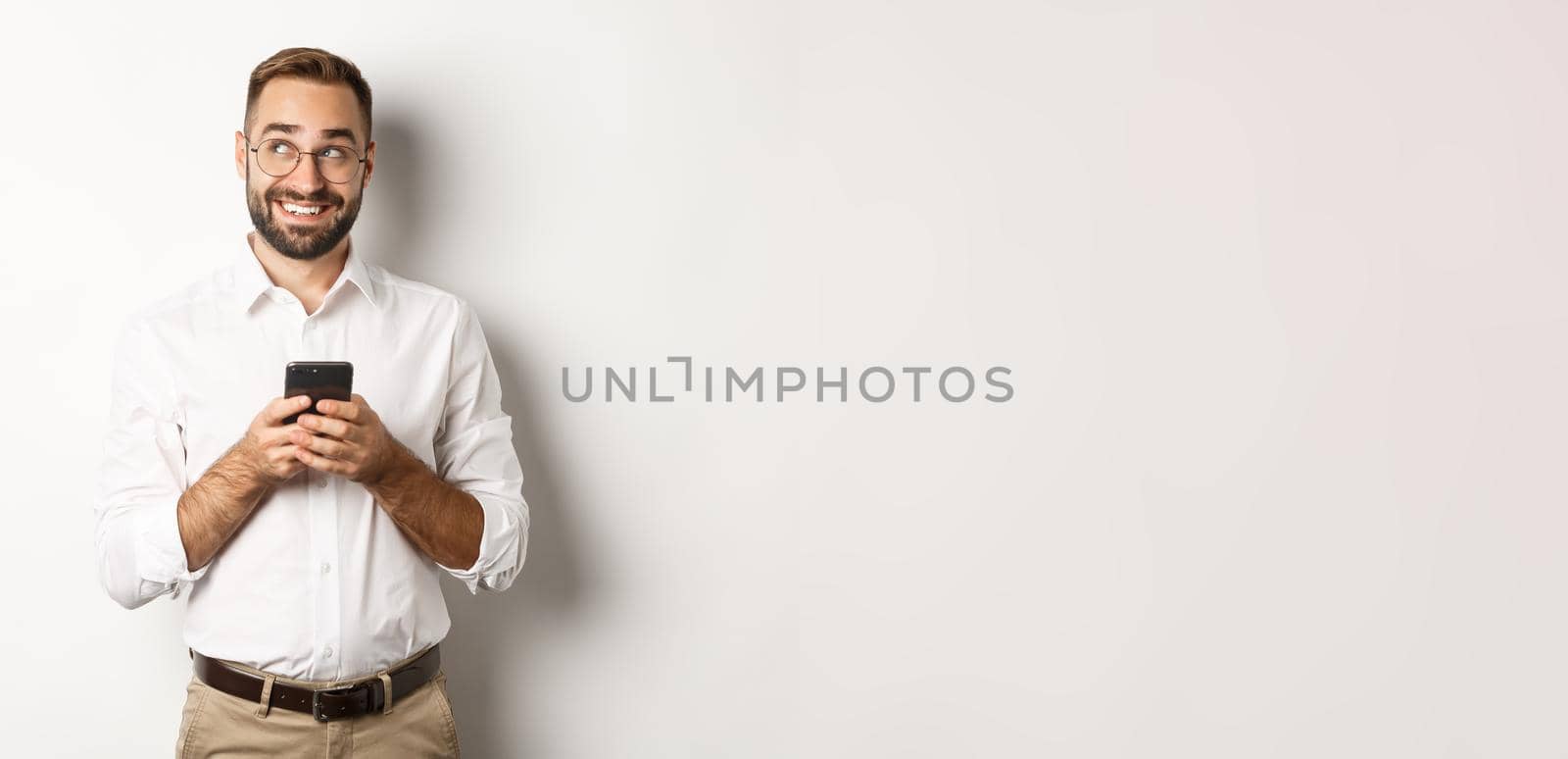 Thouthful handsome manager using mobile phone and thinking about answering message, looking at upper left corner and smiling, standing over white background.