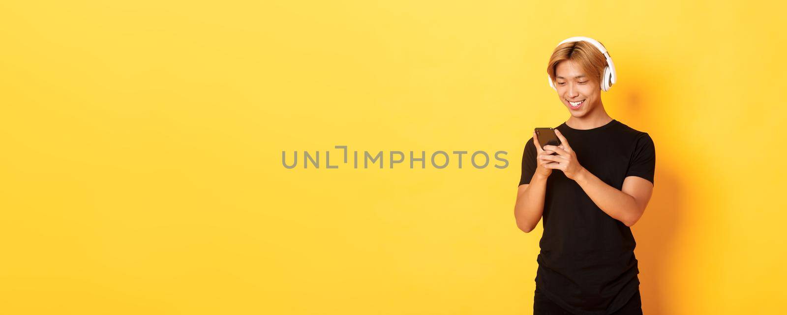 Young smiling asian guy using smartphone and listening music or podcast in wireless headphones, standing yellow background.