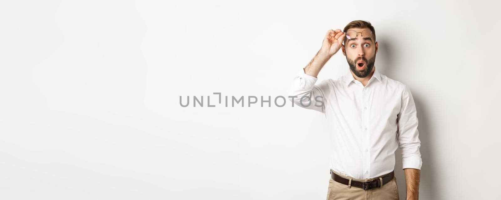 Surprised businessman taking-off his glasses, looking with amazement at camera, standing over white background.