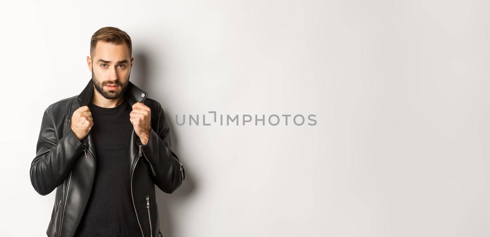 Image of handsome and confident man putting on leather biker jacket, standing against white background.