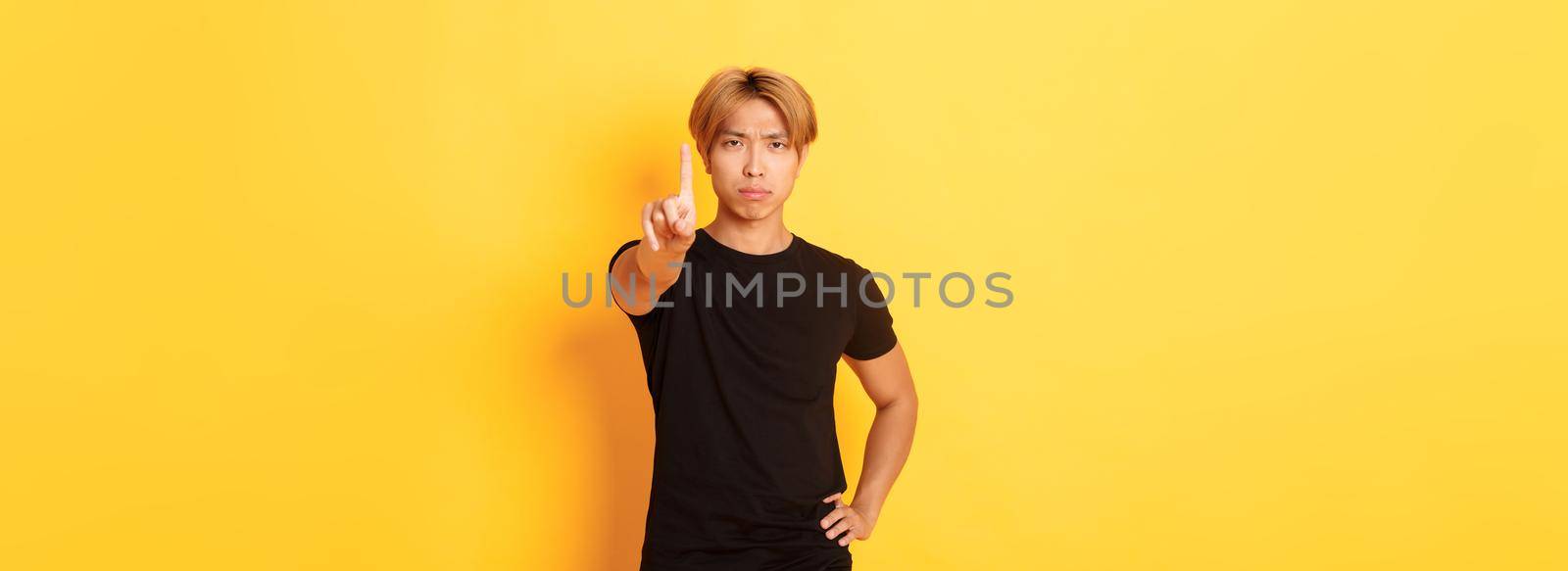 Portrait of serious-looking disappointed asian man shaking finger to scold someone, standing yellow background.