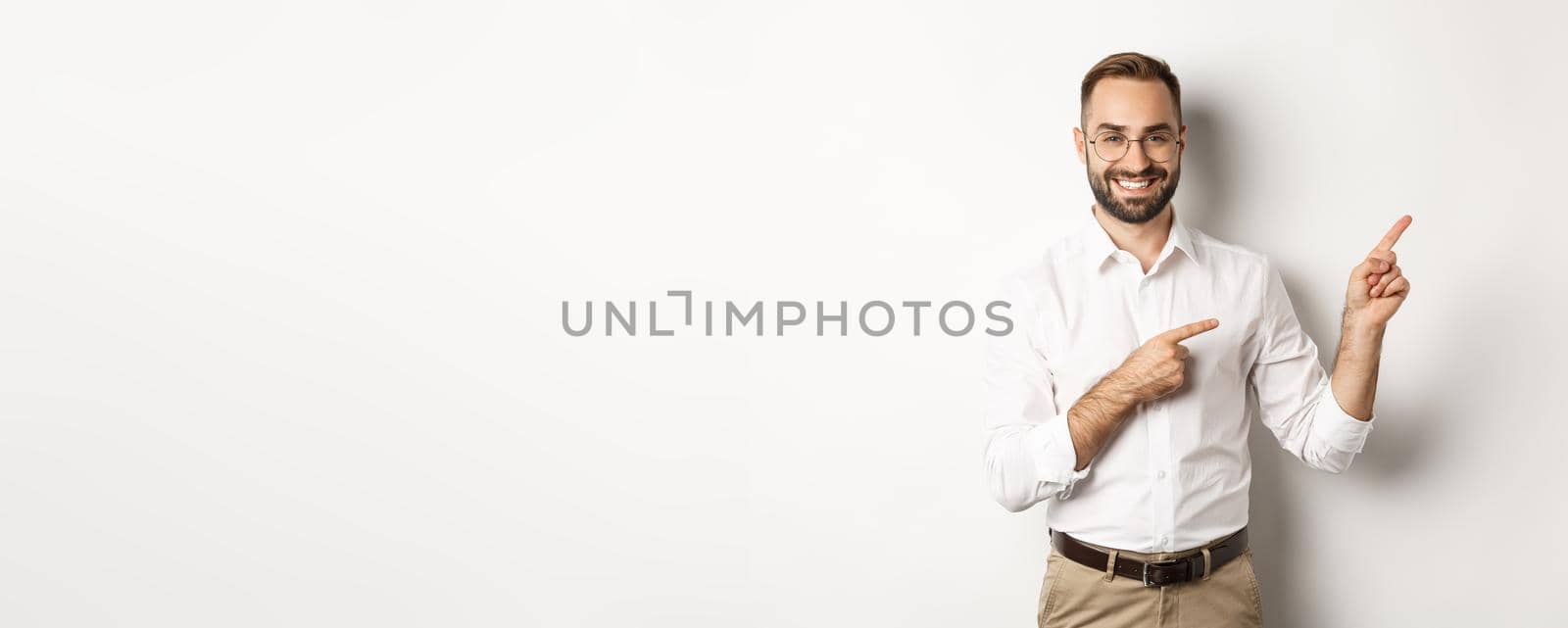 Handsome successful businessman pointing fingers right, showing advertisement with pleased face, white background.