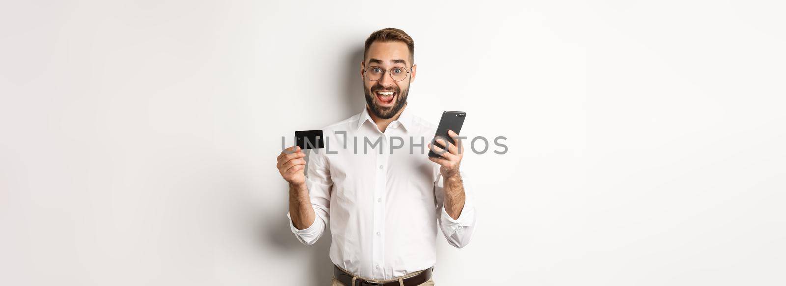 Business and online payment. Excited man paying with mobile phone and credit card, smiling amazed, standing over white background.