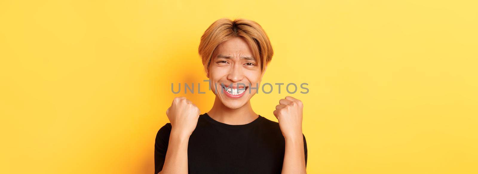 Close-up of lucky happy asian guy winning prize, making fist pump gesture and smiling satisfied, triumphing over achievement, standing yellow background.