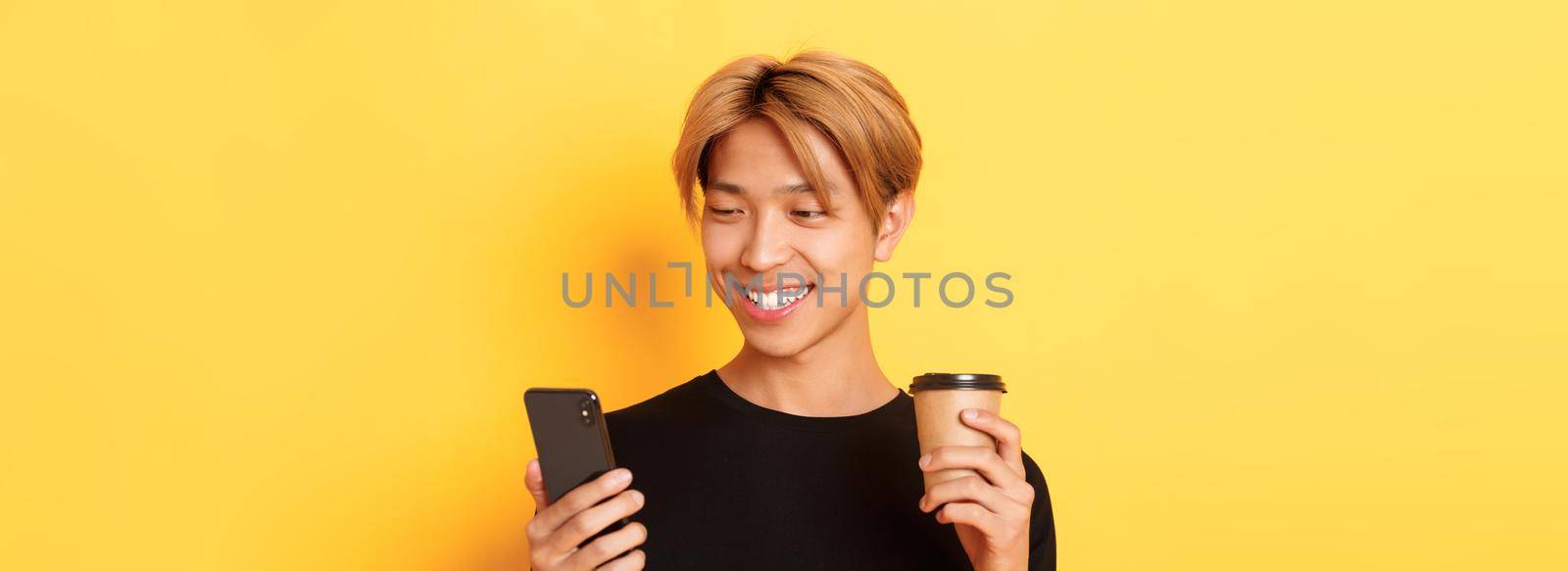 Close-up of handsome stylish asian male student, using mobile phone and drinking coffee. Guy messaging on smartphone, standing over yellow background by Benzoix