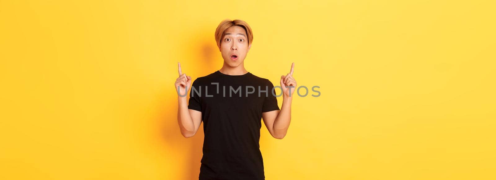 Portrait of astonished asian guy in black t-shirt, open mouth fascinated, pointing fingers up, yellow background by Benzoix