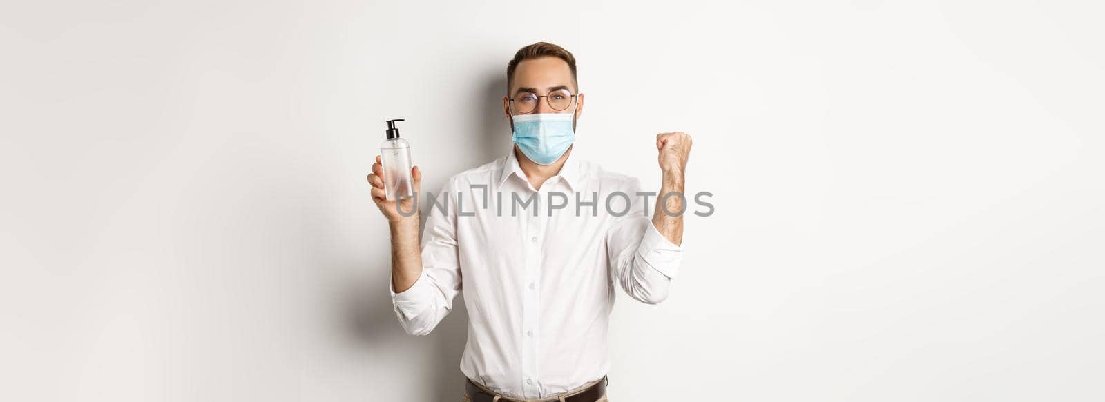 Covid-19, social distancing and quarantine concept. Cheerful manager in medical mask showing hand sanitizer, standing over white background.