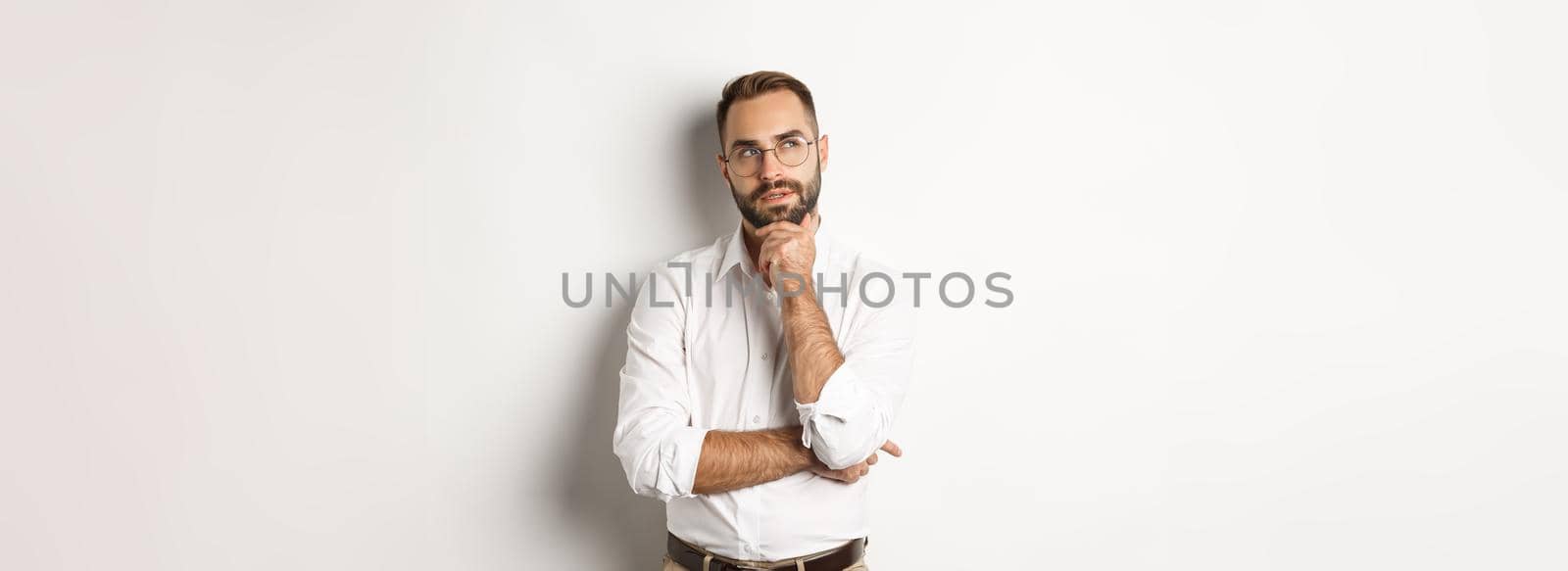 Thoughtful businessman in glasses making plan, looking at upper left corner and thinking, standing against white background.