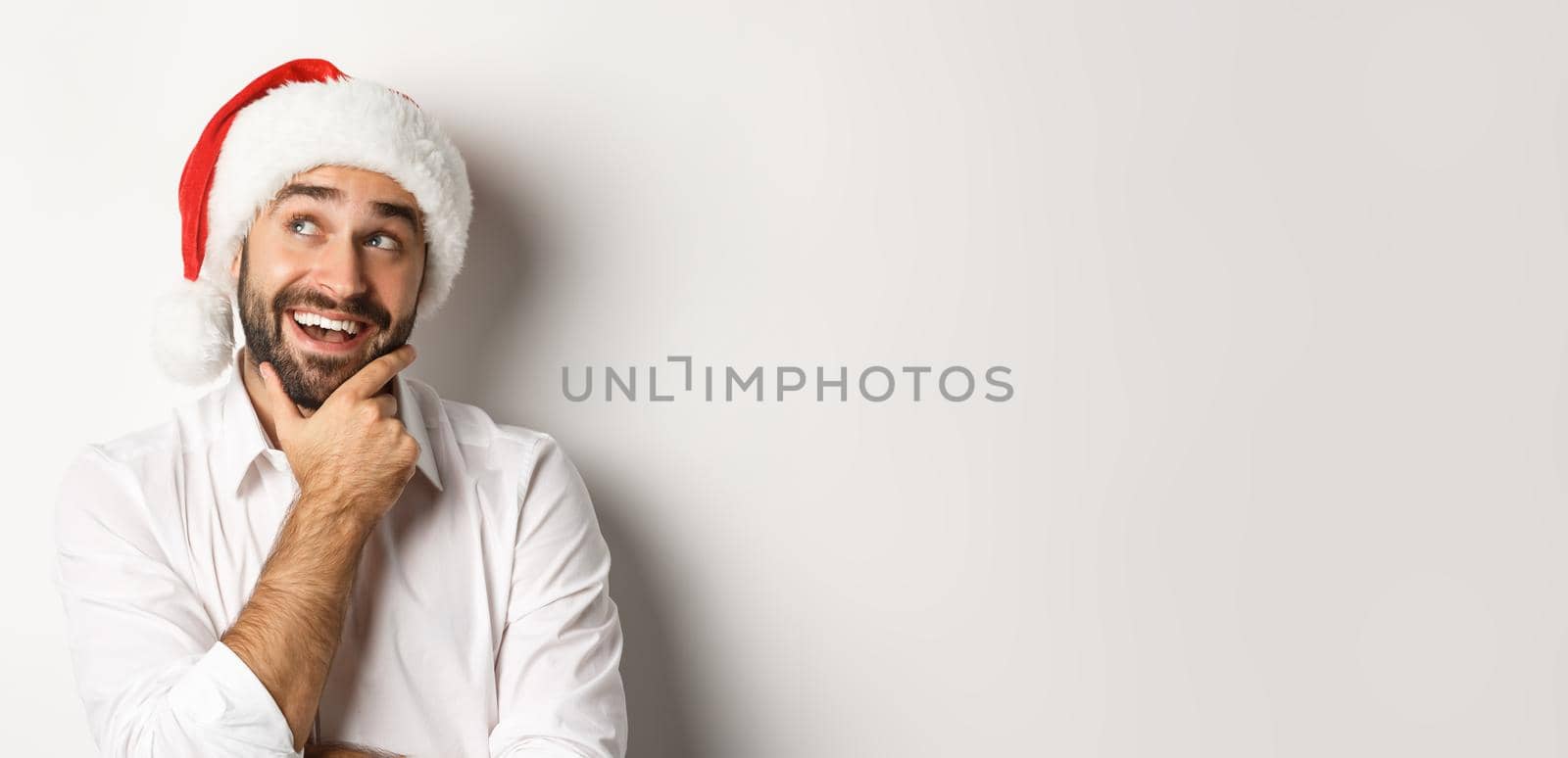 Party, winter holidays and celebration concept. Close-up of happy man planning christmas gift list, wearing santa hat, looking at upper left corner thoughtful, white background.