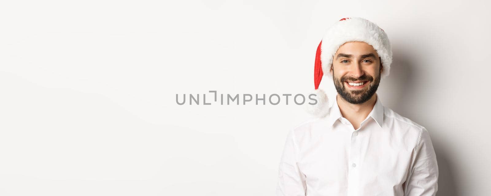 Close-up of happy bearded man celebrating christmas, wearing santa party hat and smiling, standing over white background.