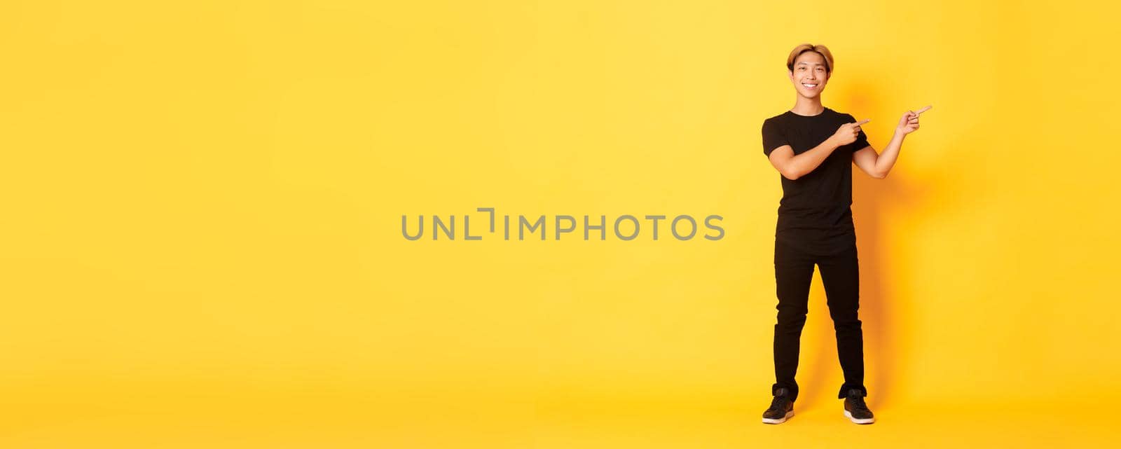 Full-length of handsome confident asian guy smiling happy, pointing fingers right, showing logo, yellow background.