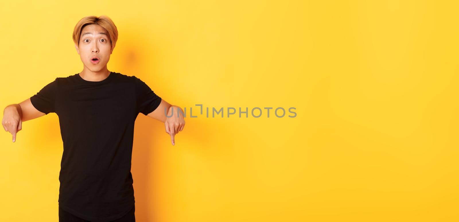 Amused happy asian guy, pointing fingers down and gasping impressed, showing awesome advertisement, yellow background by Benzoix