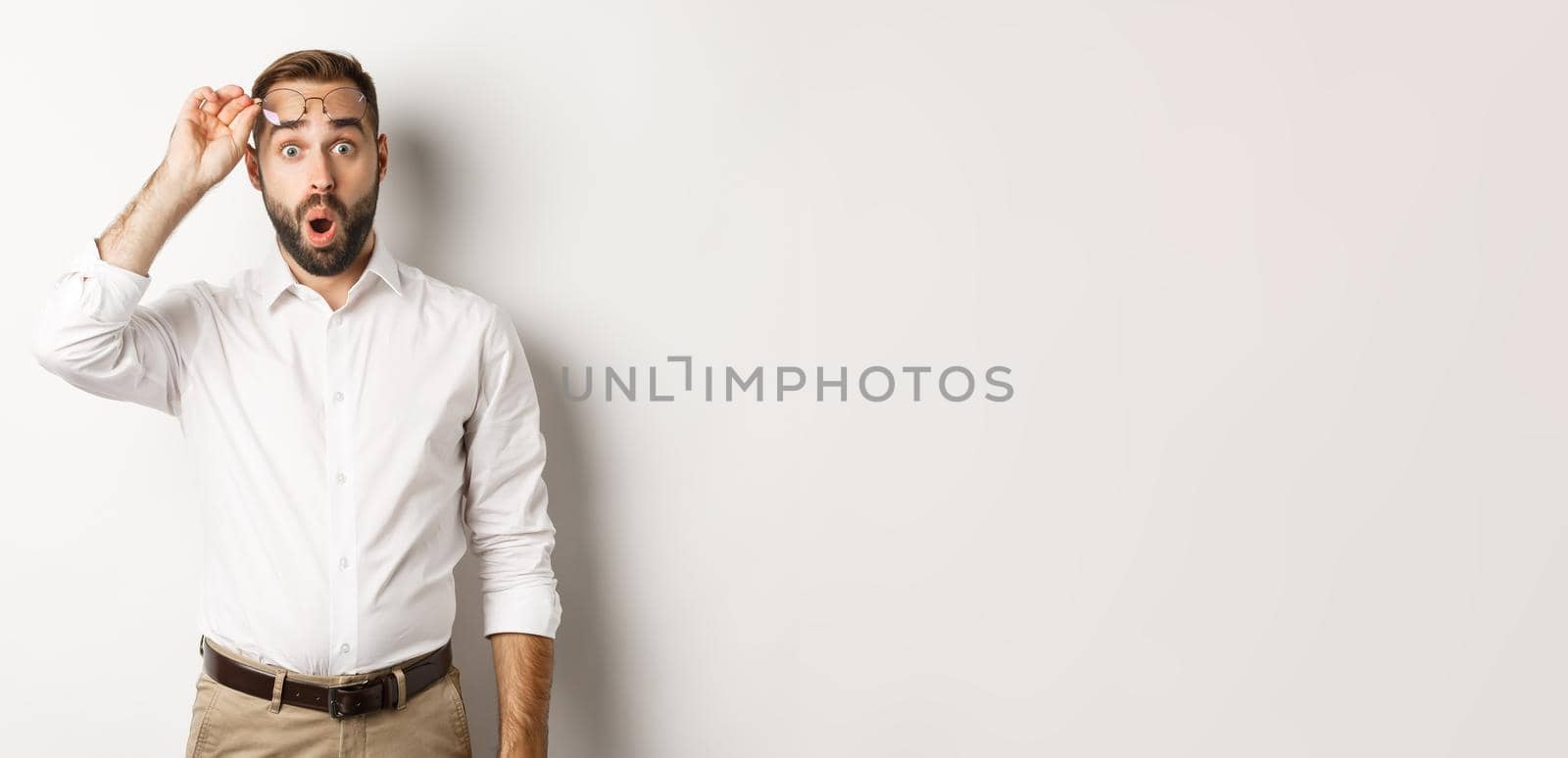 Surprised businessman taking-off his glasses, looking with amazement at camera, standing over white background.