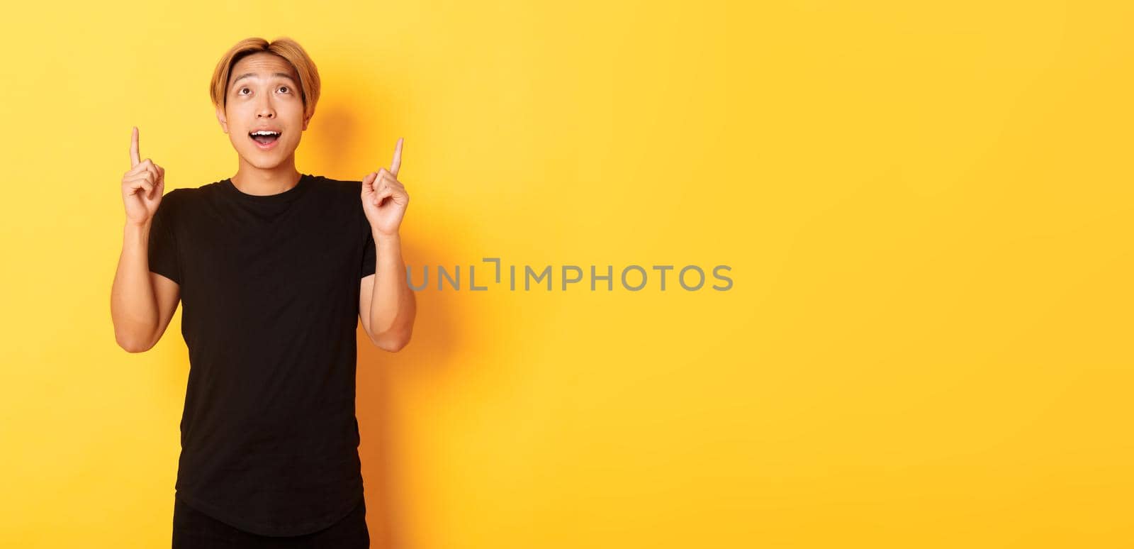 Impressed good-looking asian blond guy, wearing black t-shirt, looking and pointing fingers up excited, yellow background.