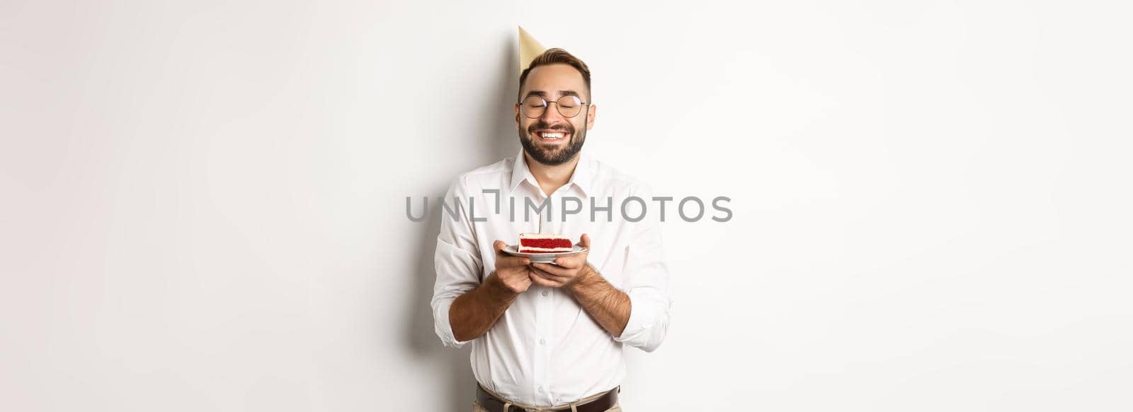 Holidays and celebration. Happy man having birthday party, making wish on b-day cake and smiling, standing against white background.