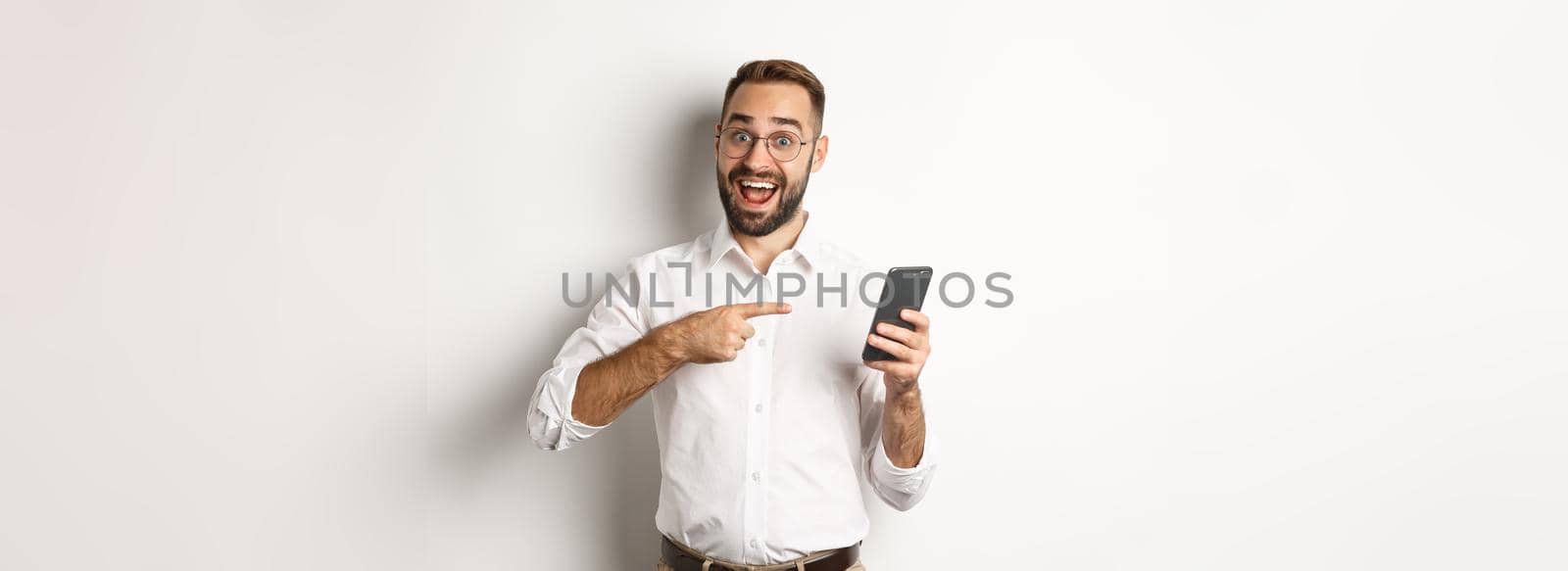 Man looking excited and pointing finger at mobile phone, showing good online offer, standing over white background.