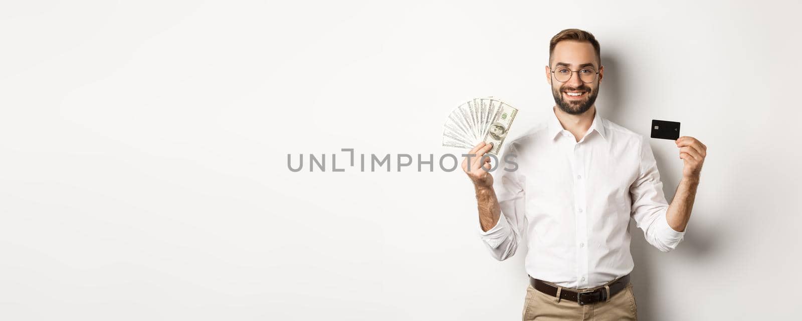 Handsome business man showing credit card and money dollars, smiling satisfied, standing over white background.