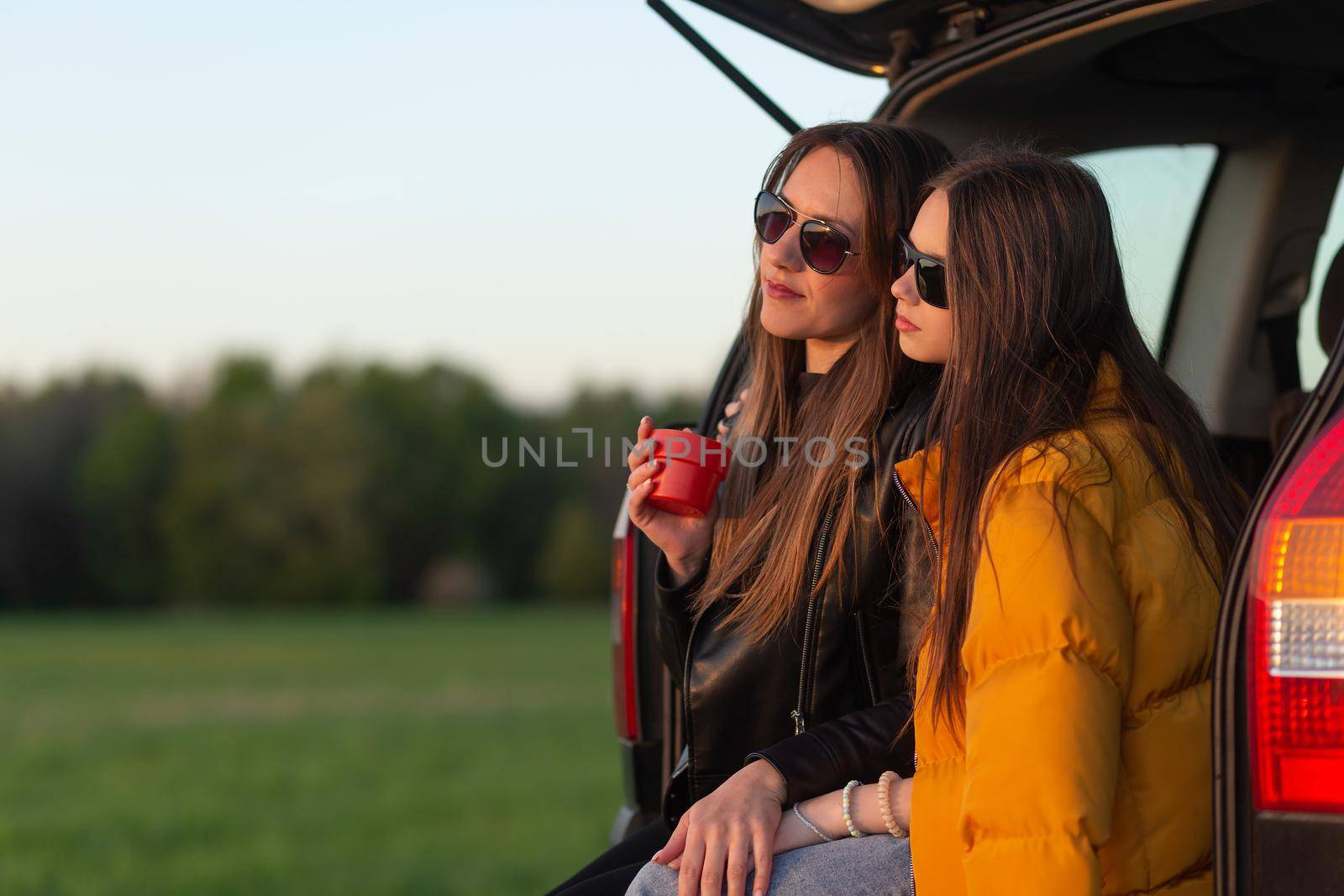 Mother and daughter camping on a hill and admiring the sunset while sitting in the car trunk